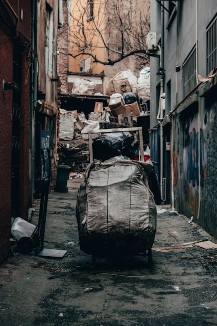 A narrow, urban alleyway cluttered with overflowing rubbish bags and discarded packaging, with a large black garbage sack positioned in the foreground on a paved surface. Behind it, a metal trolley or