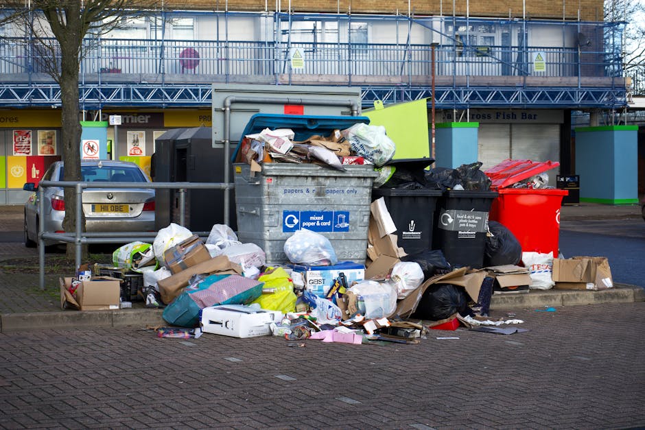 A narrow, urban alleyway cluttered with overflowing rubbish bags and discarded packaging, with a large black garbage sack positioned in the foreground on a paved surface. Behind it, a metal trolley or